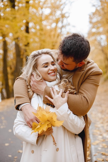 Jonge man en vrouw lopen buiten met beige jassen. Blonde vrouw en brunette man in herfst bos met gele bladeren. Romantisch paar knuffelen.