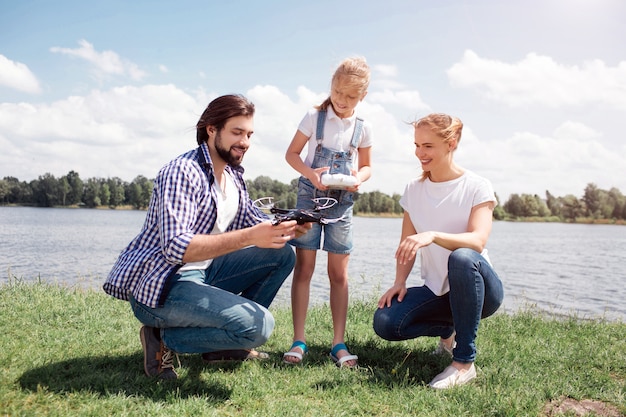 Jonge man en vrouw zitten in squat positie. Het kind staat naast hen. Guy houdt dronein handen vast. Meisje houdt bedieningspaneel. Iedereen kijkt naar drone. Ze zijn blij en glimlachen.