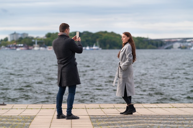 Jonge man fotografeert zijn vriendin of vrouw op een winderige promenade aan het water op een koude herfstdag met zijn mobiele telefoon