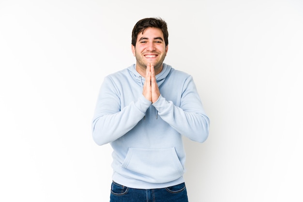 Jonge man geïsoleerd op een witte achtergrond hand in hand bidden in de buurt van de mond, voelt zich zelfverzekerd.