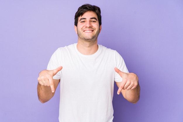 Jonge man geïsoleerd op paarse ruimte wijst naar beneden met vingers, positief gevoel.