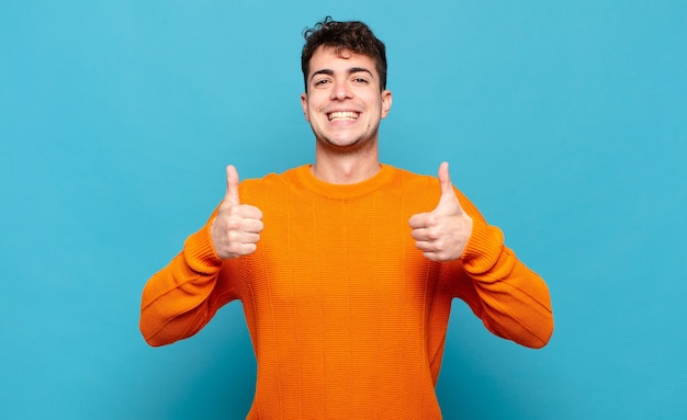 Jonge man glimlachend in brede zin gelukkig, positief, zelfverzekerd en succesvol, met beide duimen omhoog