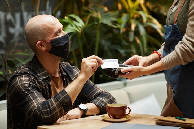 Jonge man in beschermend masker betalen voor zijn bestelling met creditcard zittend in café