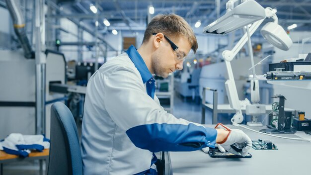 Jonge man in blauw-witte werkjas gebruikt een kleer om gedrukte printplaten te monteren voor werknemers van een smartphone-elektronicafabriek in een hightechfabriek.