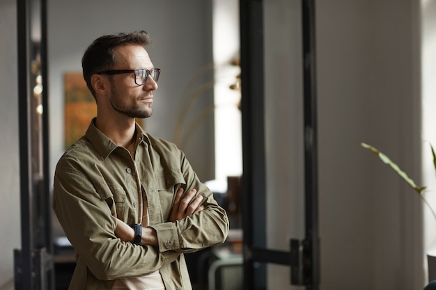 Jonge man in bril staan op kantoor en kijken door het raam met peinzende blik