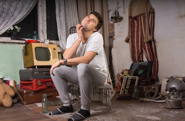 Jonge man in casual outfit die zijn nek krabt terwijl hij op de kooi in de rommelkamer zit.