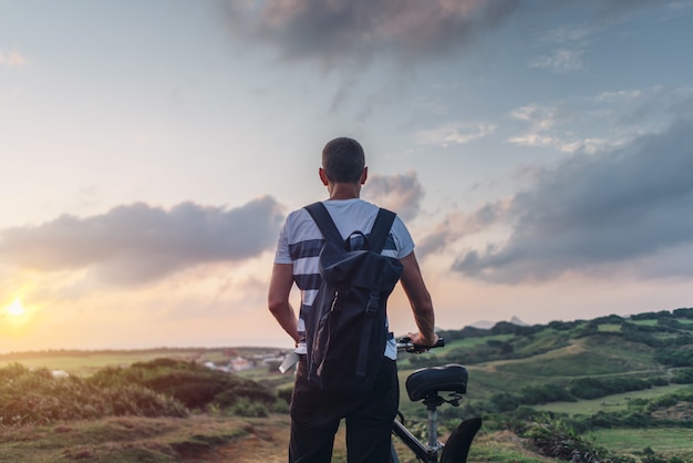 Jonge man in de avond bij zonsondergang staande op een heuvel met een fiets