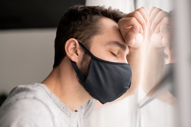 Jonge man in doek masker moe van isolatie staan met gesloten ogen voor raam, thuis in quarantaine