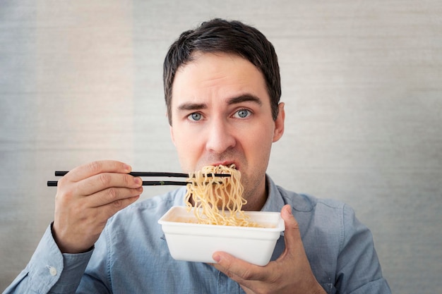 Jonge man in een blauw hemd eet noedels uit een doos met een ontevreden gezicht lunch op het kantoor