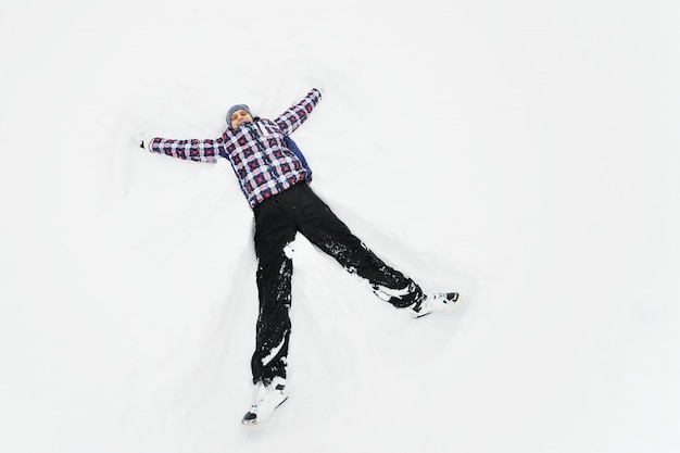 Jonge man in een geruite bovenkleding of jas ligt in de sneeuw en maakt de figuur van een engel. Winterplezier