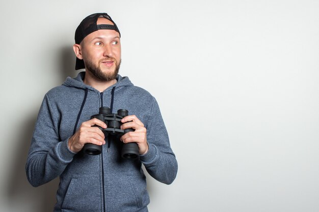 Jonge man in een hoodie en een pet met een ernstig gezicht kijkt opzij en houdt een verrekijker vast