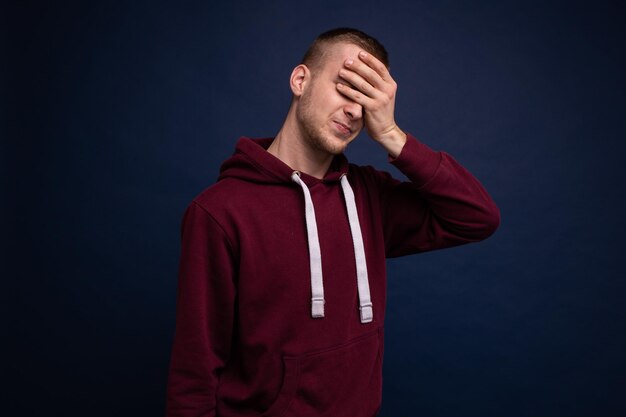 Jonge man in een rode hoodie en spijkerbroek poseren op een blauwe achtergrond, sluit zijn ogen met zijn hand