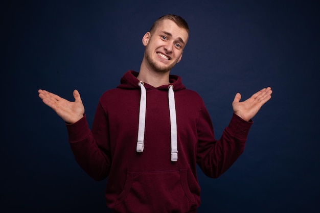 Jonge man in een rode hoodie en spijkerbroek poseren op een blauwe achtergrond, trekt een onschuldig gezicht en spreidt zijn armen naar de zijkanten