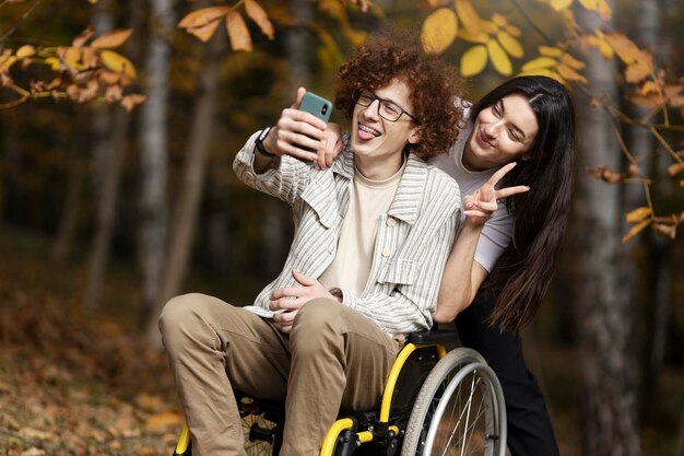 Jonge man in een rolstoel en zijn vriendin hebben plezier buitenshuis en maken een selfie Jonge mensen trekken grappige gezichten