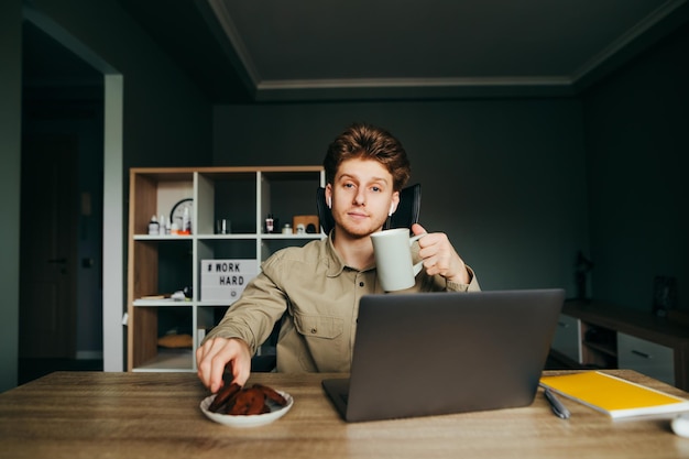 jonge man in een shirt en draadloze koptelefoon die thuis op het werk zit en op een laptop werkt