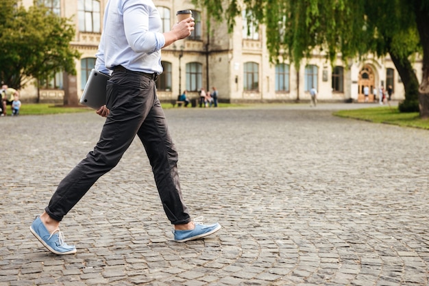 Foto jonge man in formele slijtage, wandelen door stadsstraat met afhaalmaaltijden koffie en zilveren laptop