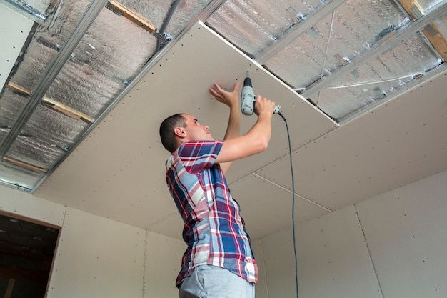 Foto jonge man in gebruikelijke kleding en werkhandschoenen tot vaststelling van gipsplaten verlaagd plafond aan metalen frame met behulp van elektrische schroevendraaier op plafond geïsoleerd met glanzende aluminiumfolie. diy, doe het zelf concept.