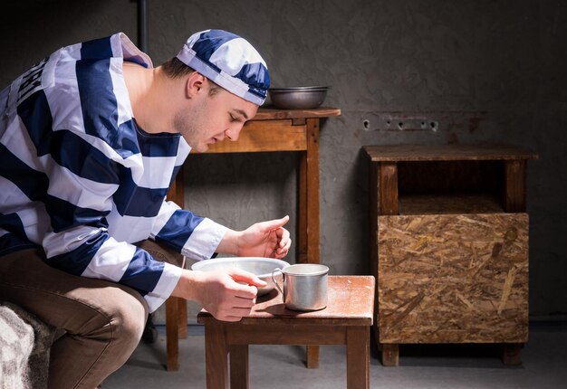 Jonge man in gevangenisuniform zittend op een bed en etend van aluminium schotels in een kleine gevangeniscel