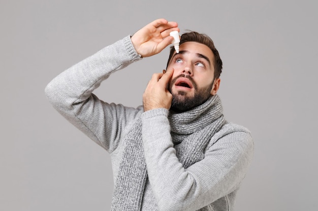 Jonge man in grijze trui, sjaal geïsoleerd op grijze muur achtergrond studio portret. Gezonde levensstijl, behandeling van zieke ziektes, concept van het koude seizoen. Bespotten kopie ruimte. Vasthouden, druipende oogdruppels.