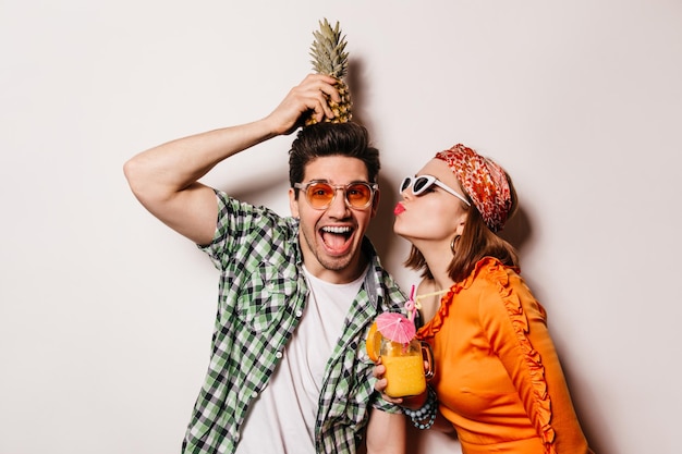 Jonge man in groen shirt en wit t-shirt houdt ananas op zijn hoofd meisje in oranje jurk met cocktail in haar handen kust man op wang