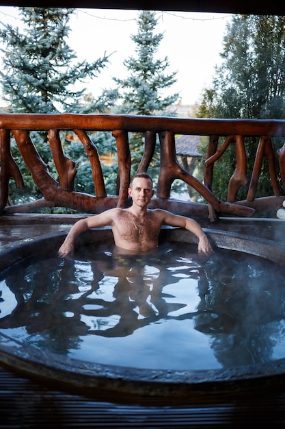 Jonge man in het zwembad met warm water op het terras, lichaamsbehandelingen in de spa. Buitenjacuzzi. Jacuzzi met een man. Ontspan in de open lucht. Huisje met bubbelbad.
