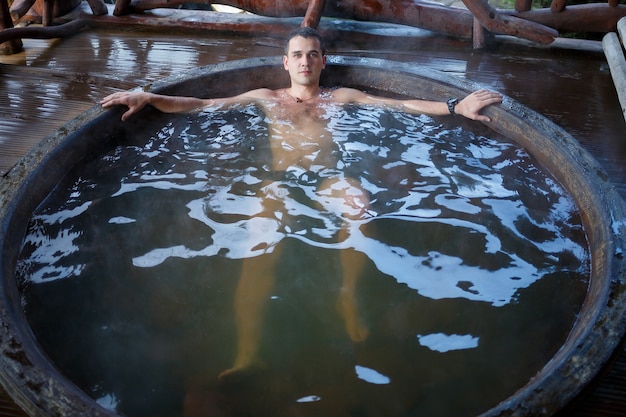 Jonge man in het zwembad met warm water op het terras, lichaamsbehandelingen in de spa. Buitenjacuzzi. Jacuzzi met een man. Ontspan in de open lucht. Huisje met bubbelbad.