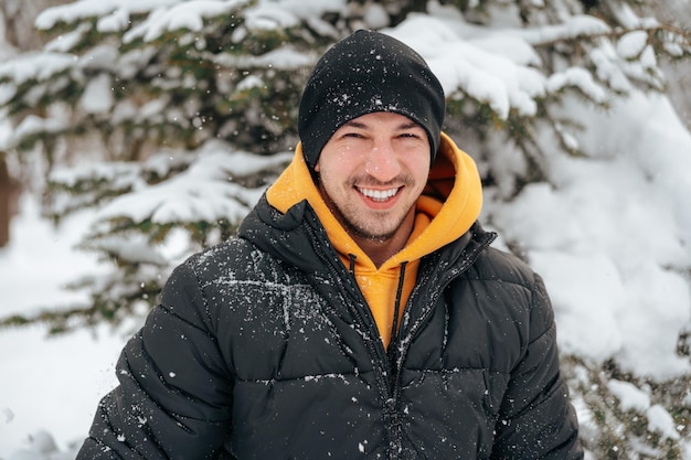 Jonge man in hoodie en warme jas die in een besneeuwd park staat en glimlacht