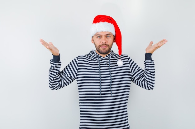 Jonge man in hoodie, kerstman hoed spreidt handpalmen zijwaarts en ziet er zelfverzekerd uit, vooraanzicht.