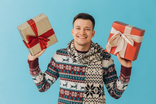 Jonge man in kersttrui en sjaal met veel geschenkdozen met cadeau lint strik geïsoleerd op blauwe achtergrond Gelukkig nieuwjaar viering concept