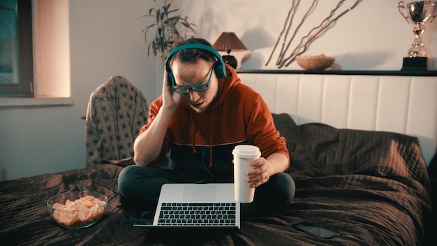 Jonge man in koptelefoon zittend op het bed en praten met iemand met behulp van laptop