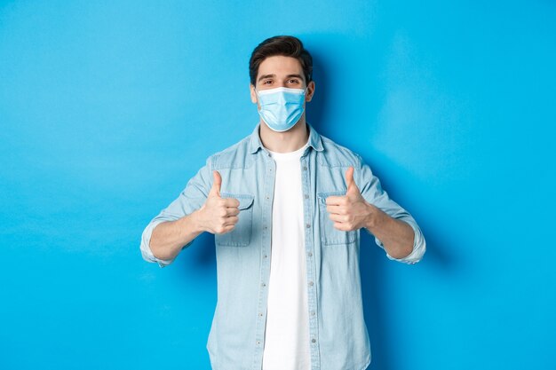 Jonge man in medisch masker met thumbs-up in goedkeuring, zoals en ga akkoord, staande tegen blauwe muur