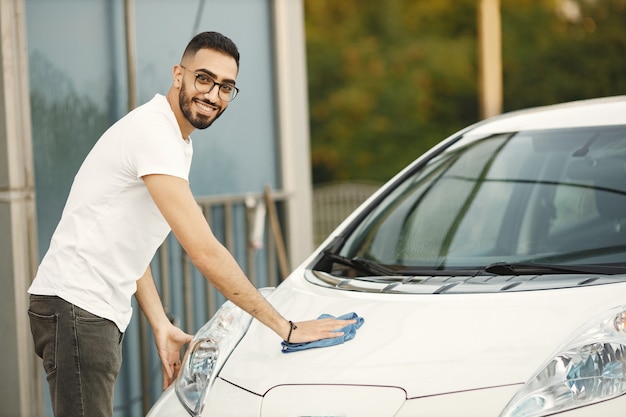 Jonge man in modekleding die zijn auto afveegt met een kleed na het wassen bij het autowasstation. Man met wit t-shirt en spijkerbroek
