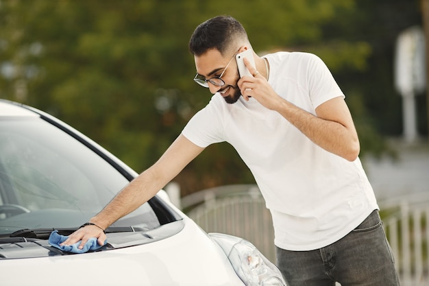 Jonge man in modekleding die zijn auto afveegt met een kleed na het wassen bij het autowasstation. Man met wit t-shirt en spijkerbroek