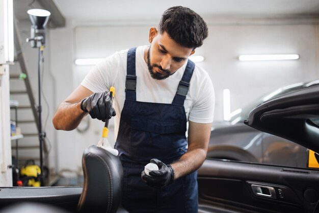 Jonge man in overall en beschermende handschoenen autostoeltje schoonmaken met zachte borstel en schuim