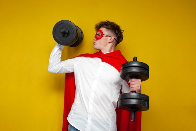 Jonge man in superman kostuum tillen zware dumbbells en doet sport op gele achtergrond