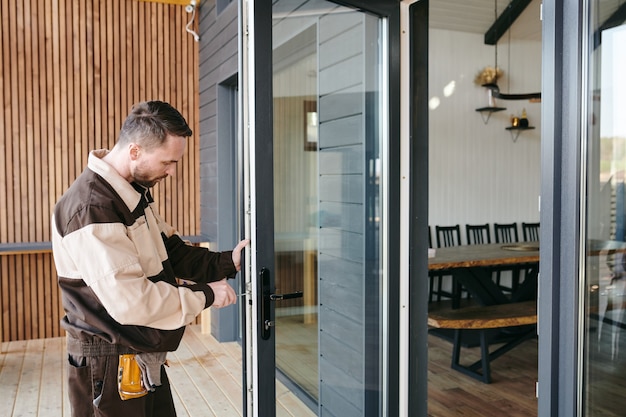 Jonge man in uniform met handgereedschap tijdens het repareren van de deur van een groot modern huis