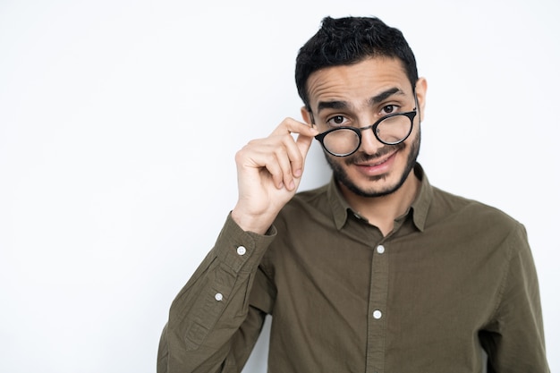 Jonge man in vrijetijdskleding die zijn bril opstijgt terwijl hij u geïsoleerd tegen witte ruimte bekijkt