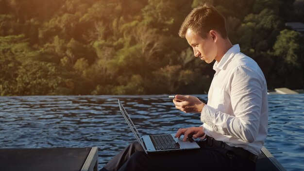 Foto jonge man in wit overhemd werkt op laptop en kijkt naar de telefoon tegen groene planten en blauw zwembadwater bij zonsondergang