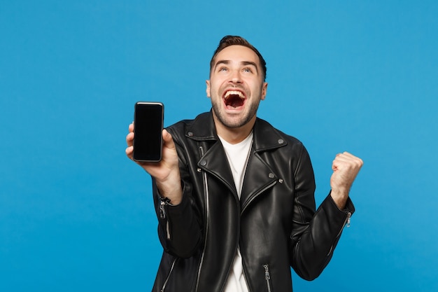 Jonge man in zwarte jas wit t-shirt houdt mobiele telefoon met leeg leeg scherm voor promotionele inhoud geïsoleerd op blauwe muur achtergrond studio portret. Mensen levensstijl concept. Bespotten kopie ruimte