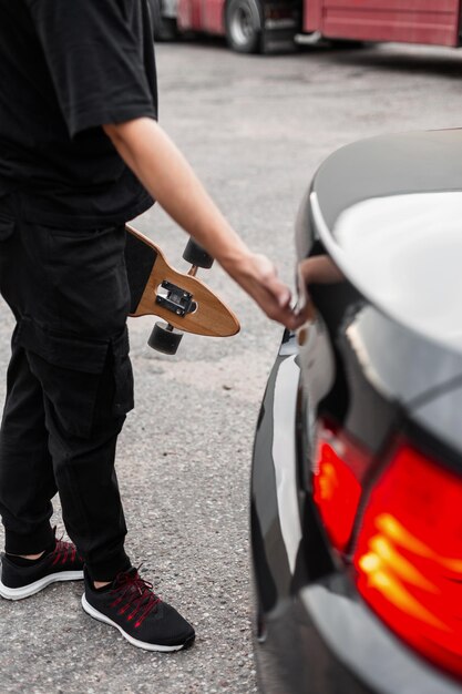 Jonge man in zwarte kleding met longboarden opent de kofferbak van een zwarte auto Actief weekendconcept