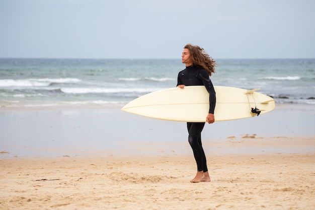 Jonge man in zwarte wetsuit wandelen in het strand met surfplank overdag