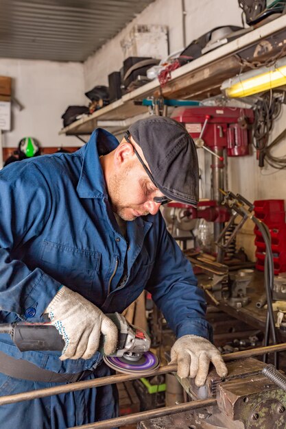 jonge man lasser in een blauw T-shirt, bril en bouwhandschoenen verwerkt metaal een haakse slijper in de garage