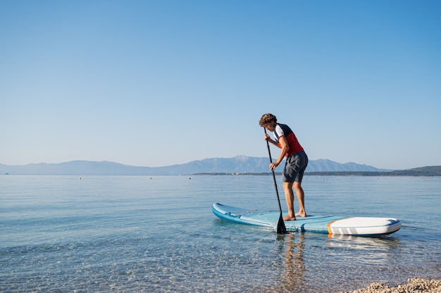 Jonge man leert peddelen op een sup board drijvend op kalm ochtend zeewater.