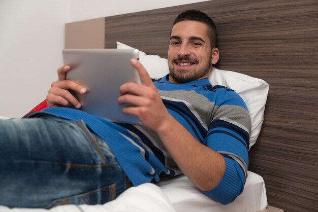 Jonge man liggend op bed en plezier met touchpad in slaapkamer