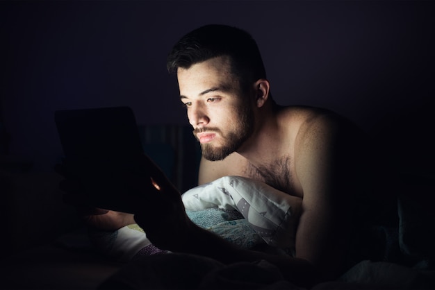 Jonge man liggend op bed op zijn buik en kijk naar het scherm van de tablet. Geconcentreerde kalme man die spelletjes speelt of zich vermaakt. Donker 's nachts.