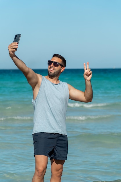 Jonge man met baard en zonnebril die op een zonnige dag een selfie maakt met zijn smartphone op het strand