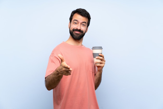 Jonge man met baard met een take-away koffie over geïsoleerde blauwe muur handen schudden voor het sluiten van een goede deal