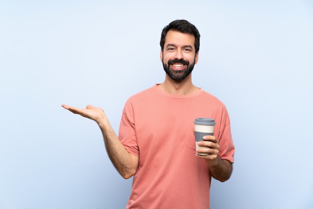 Jonge man met baard met een take-away koffie over geïsoleerde blauwe muur met copyspace denkbeeldig op de palm om een advertentie in te voegen