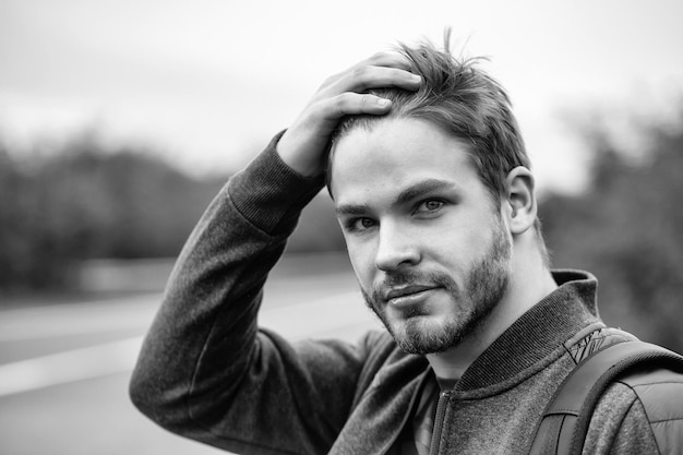 Jonge man met baard op charmante knappe sexy serieuze gezicht blauwe ogen in casual kleding hand op zijn hoofd leggen poseren op natuurlijke weg achtergrond buiten