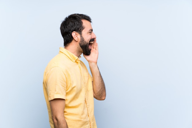 Jonge man met baard over geïsoleerde blauwe muur schreeuwen met mond wijd open voor de zijtak